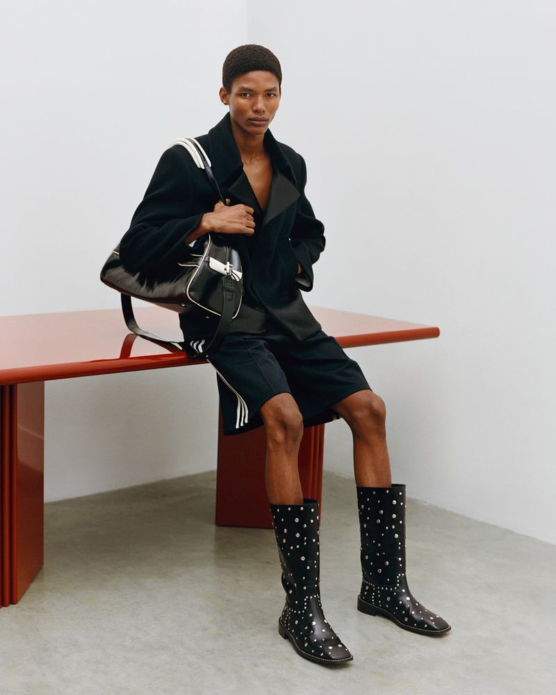 Fashionable adult in a black double-breasted coat and black shorts poses indoors next to a red table while holding a black purse.