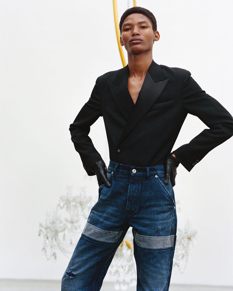 Fashionable Black male model poses indoors wearing a black blazer, dark denim jeans with patches, and black gloves. Chandelier backdrop.
