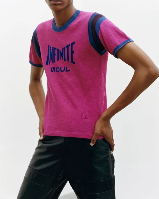 Close-up of a fashion model in a pink T-shirt with blue trim and black pants against a white backdrop, showcasing a casual yet stylish look.