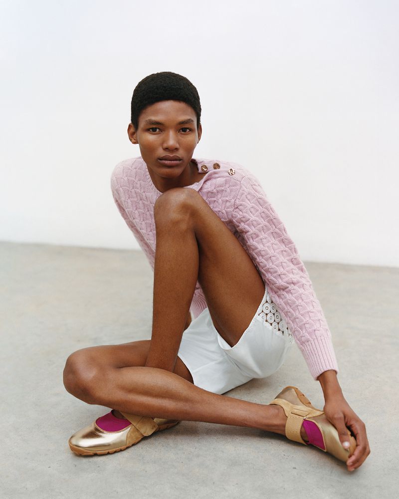 Model seated on the floor wearing a pink textured knit sweater, white shorts, and metallic gold shoes against a plain white background.