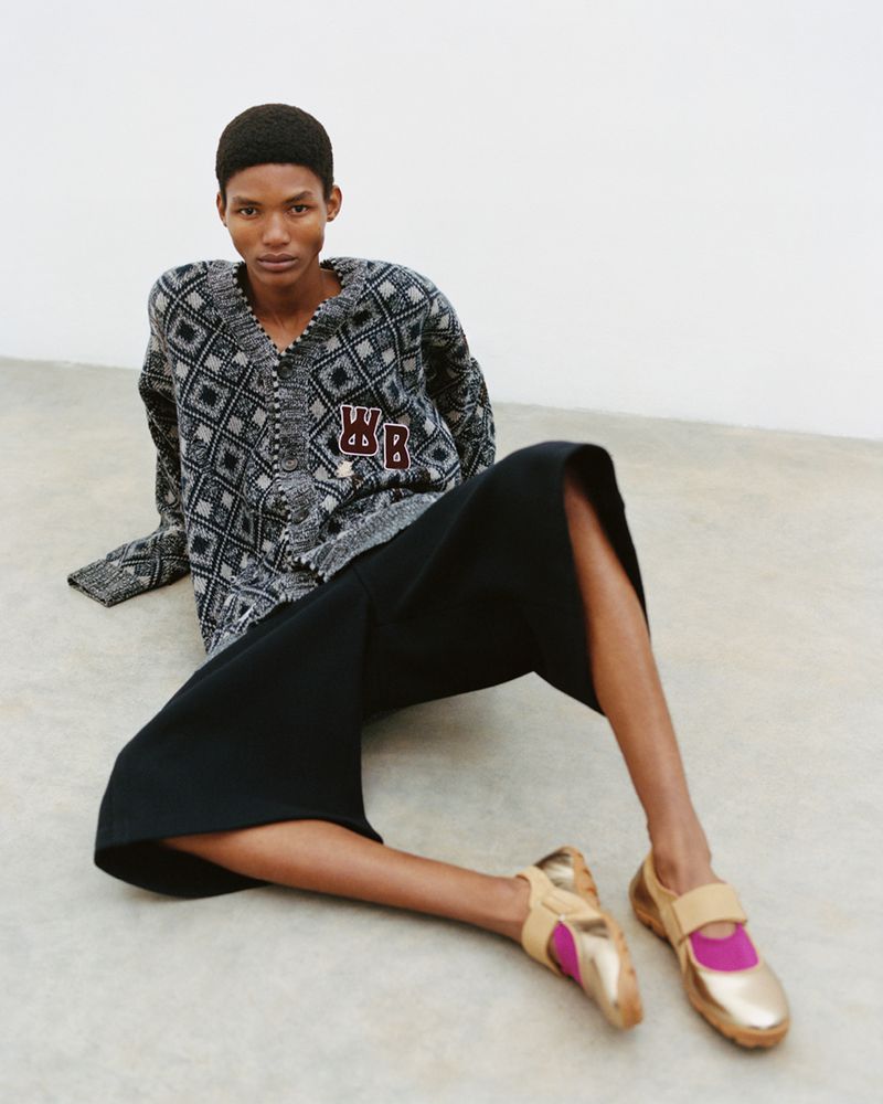 Woman sits on the floor wearing a patterned cardigan, black culottes, and gold-and-pink Mary Jane style shoes in a fashion shot.
