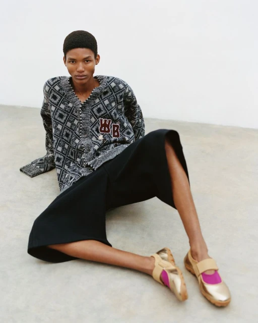 Woman sits on the floor wearing a patterned cardigan, black culottes, and gold-and-pink Mary Jane style shoes in a fashion shot.