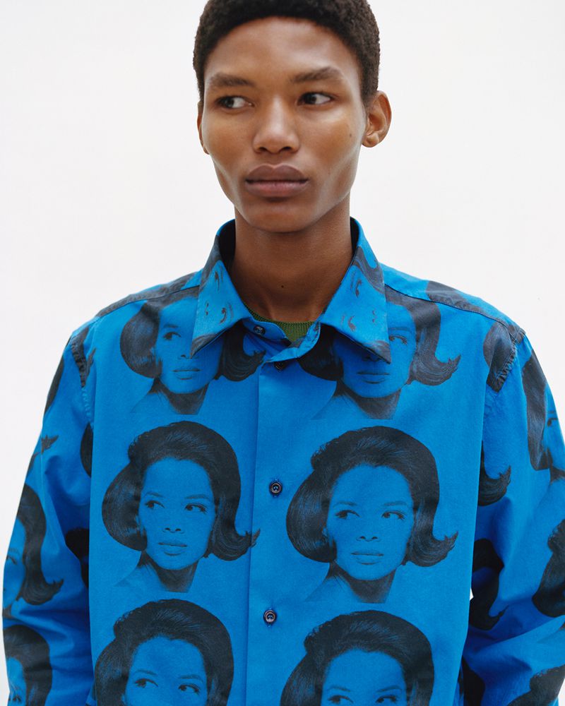 Dark-skinned man in a bright blue long-sleeve button-down shirt with black-and-white portrait graphics, standing against a white backdrop.