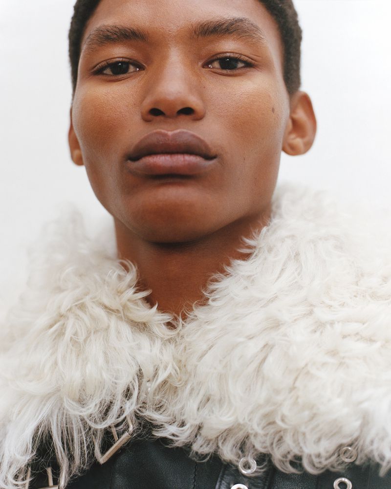 Portrait of a Black male model wearing a black leather jacket with a white shearling collar against a simple white backdrop.