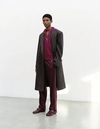 Model wearing a dark wool coat, purple zip-up jacket, and matching trousers against a plain white studio wall.