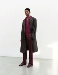Model wearing a dark wool coat, purple zip-up jacket, and matching trousers against a plain white studio wall.
