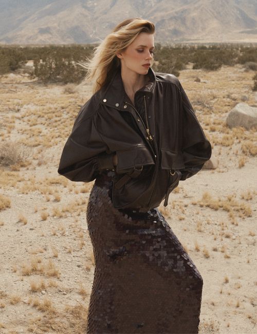 Model in a dark leather jacket and sequined skirt standing outdoors in a desert landscape with sparse bushes under a hazy sky.