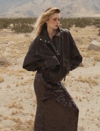 Model in a dark leather jacket and sequined skirt standing outdoors in a desert landscape with sparse bushes under a hazy sky.