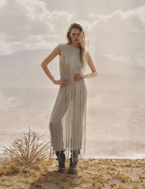 A blonde woman posing outdoors in a fringe dress and boots against a hazy mountain and desert landscape.