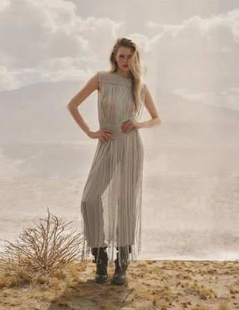 A blonde woman posing outdoors in a fringe dress and boots against a hazy mountain and desert landscape.