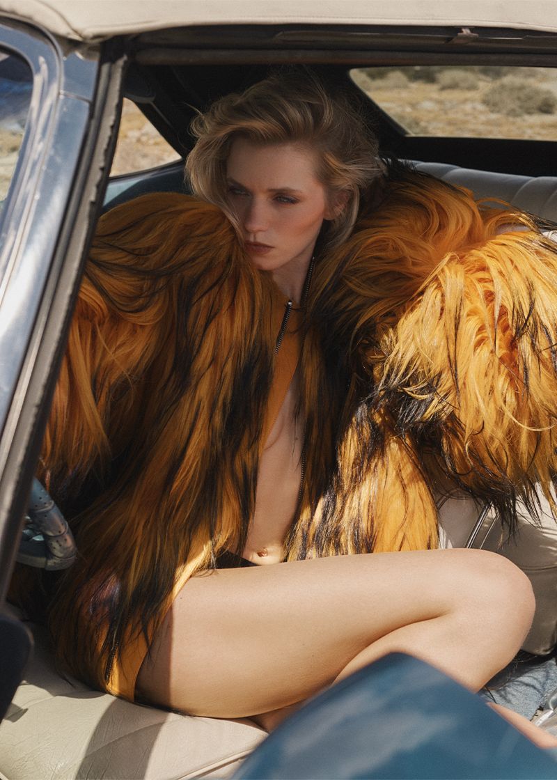 Blonde woman in a large fur coat sits inside a convertible car with tan desert environment visible in the background on a sunny day.
