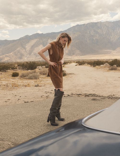 A blonde woman in a brown dress poses in the desert near mountains. She wears high boots and the front of a black car is visible.