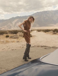 Model standing outdoors in a desert landscape wearing a brown dress and tall boots, near a car hood