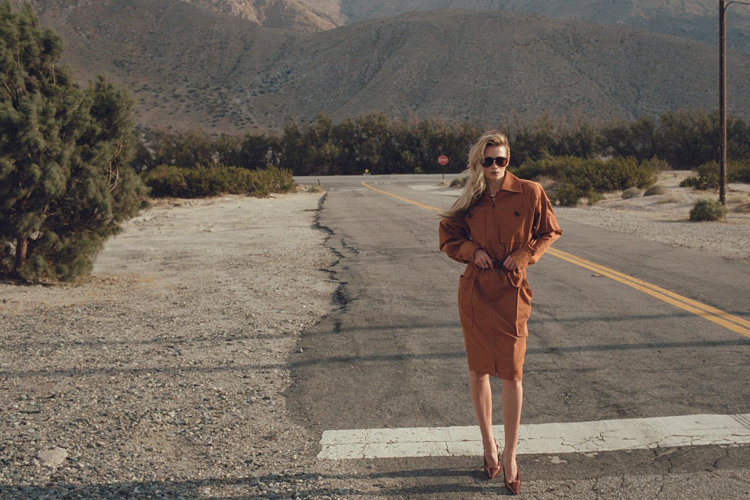 A blonde woman in sunglasses stands on an empty desert road wearing a brown shirt dress and heels with mountains in the background.