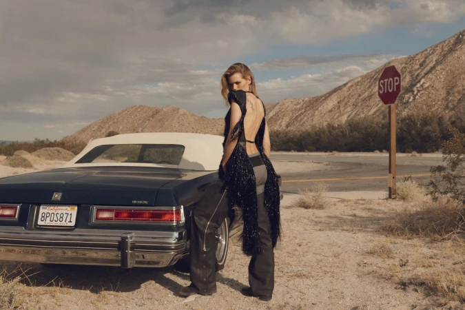 Adult woman in an open back top and wide leg pants stands next to a blue convertible in a desert landscape under a cloudy sky.