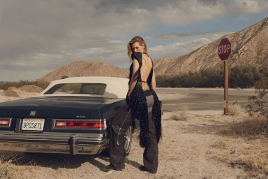 Model standing by a vintage car in a desert landscape wearing a black fringed top and dark trousers.