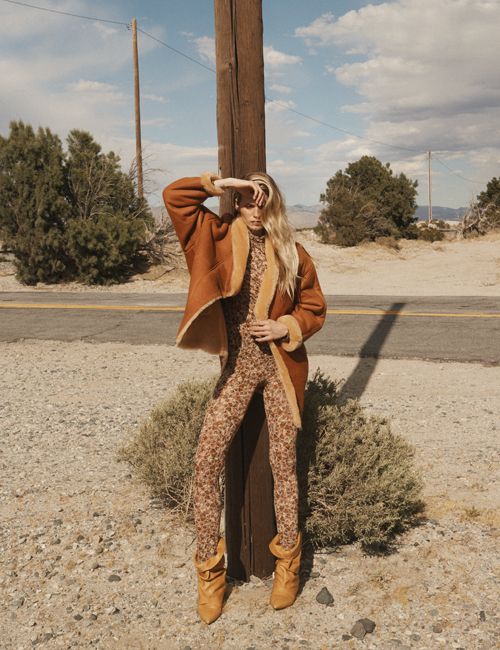A woman stands on a rural road in a shearling coat and floral-print jumpsuit, hand shielding her eyes, next to a utility pole in desert scenery.