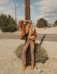 Model in patterned jumpsuit and brown shearling jacket leaning against a wooden utility pole outdoors