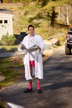 Man in bathrobe and pink shorts stands on driveway, reading a magazine. House, trees, and truck are visible in the background.