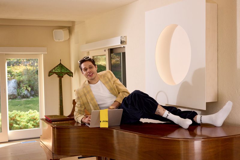 Adult male with sunglasses on head lying on a piano with a laptop. Green lawn visible through a sliding glass door behind him.