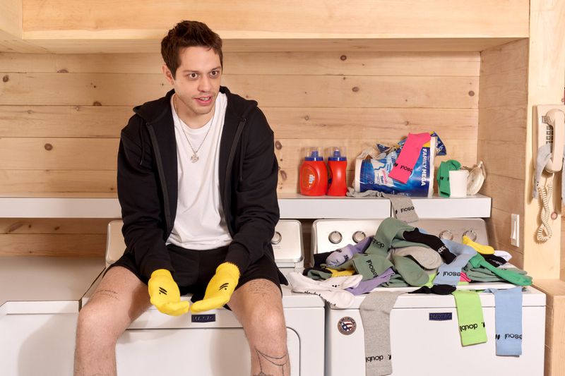 Adult male sitting on a washing machine with yellow socks for gloves, socks on top of the machine, and wood paneling in the background.