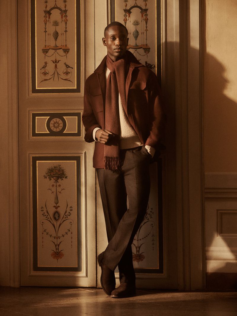 A dark-skinned man in a brown jacket and scarf poses against an ornate painted door in a warmly lit indoor fashion shot.