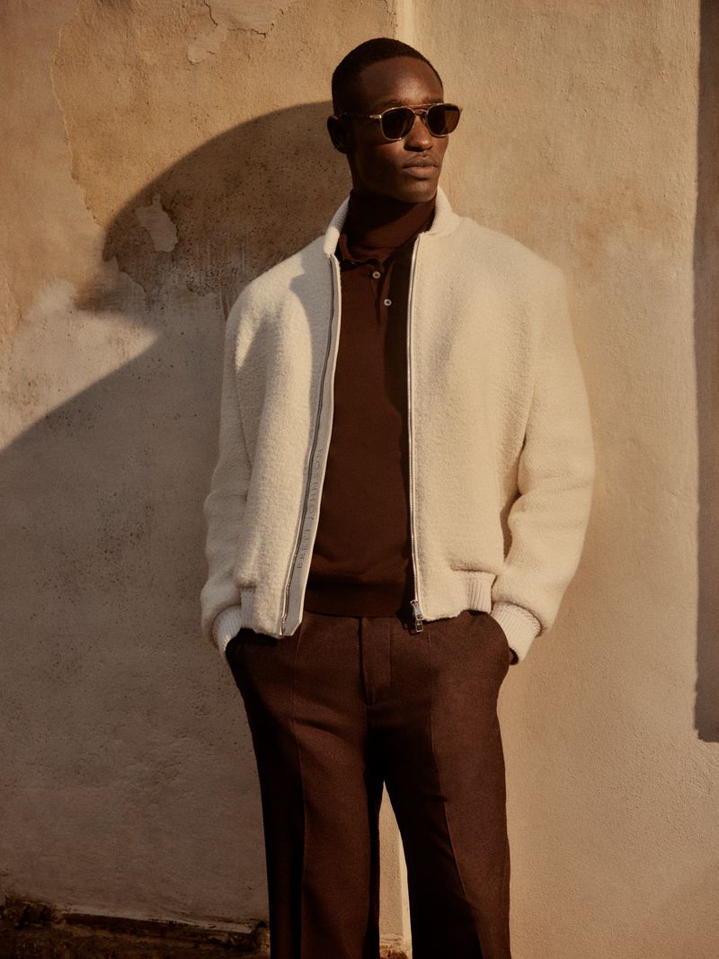 A stylish adult male wearing sunglasses, a white textured jacket, and brown pants poses in a studio with a textured wall backdrop.