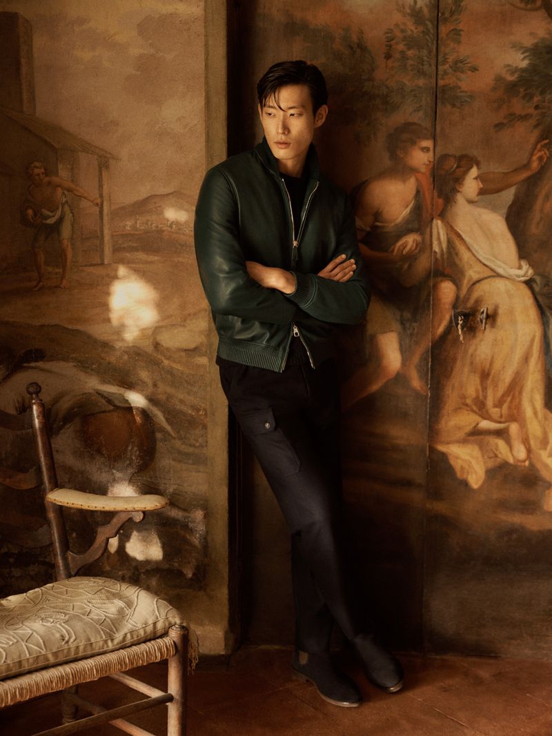 Stylish Asian male model in green jacket poses against a painterly backdrop with a chair. Fashion editorial style and indoor setting.