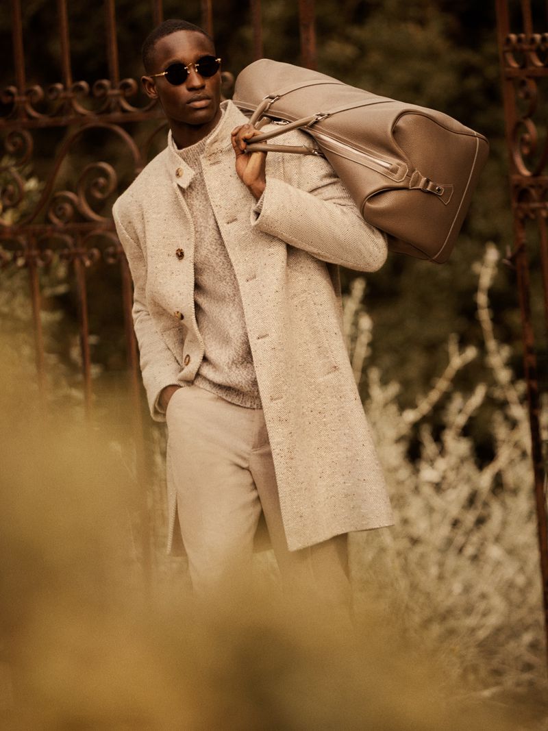 Elegant African American man in neutral-toned coat, sweater, and trousers carries leather duffel bag outdoors, styled with sunglasses.