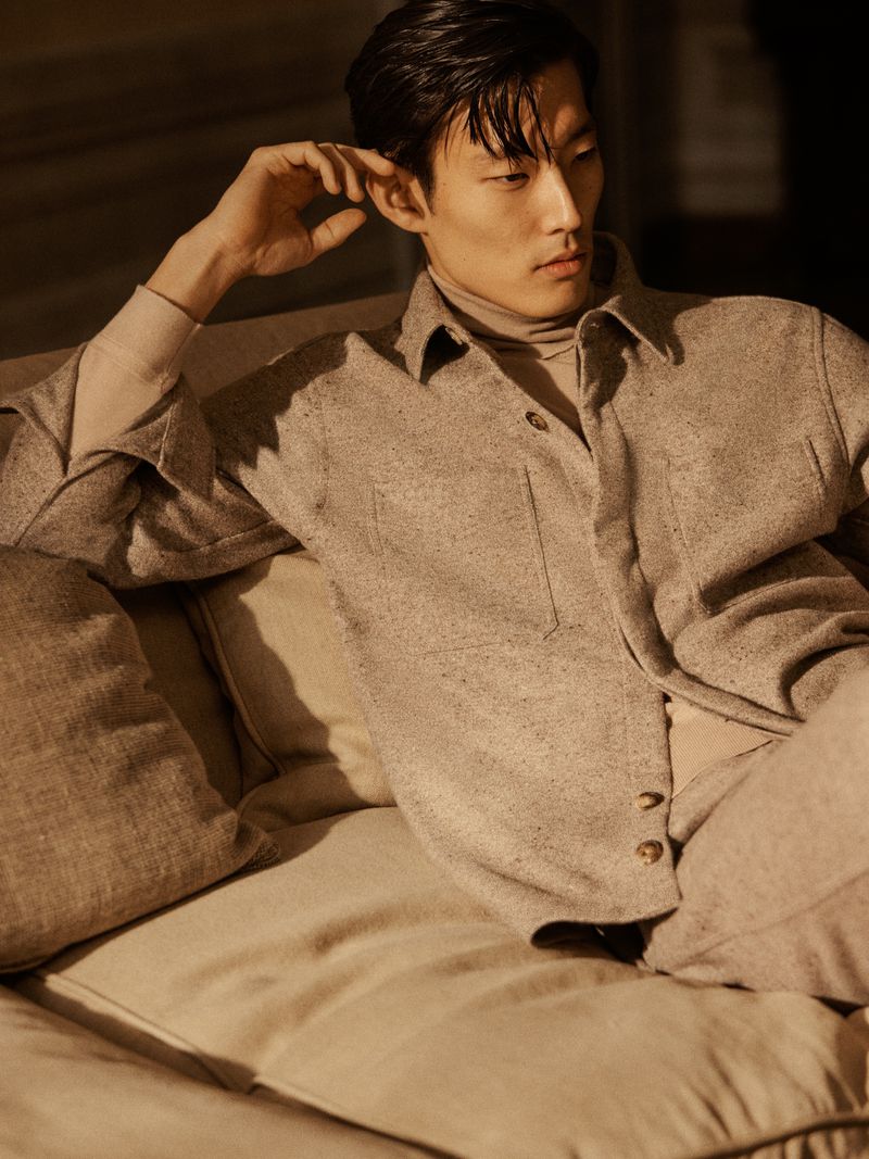 A relaxed Asian man is lounging on a sofa in a stylish neutral-toned outfit, bathed in warm, soft lighting. Relaxed and stylish.