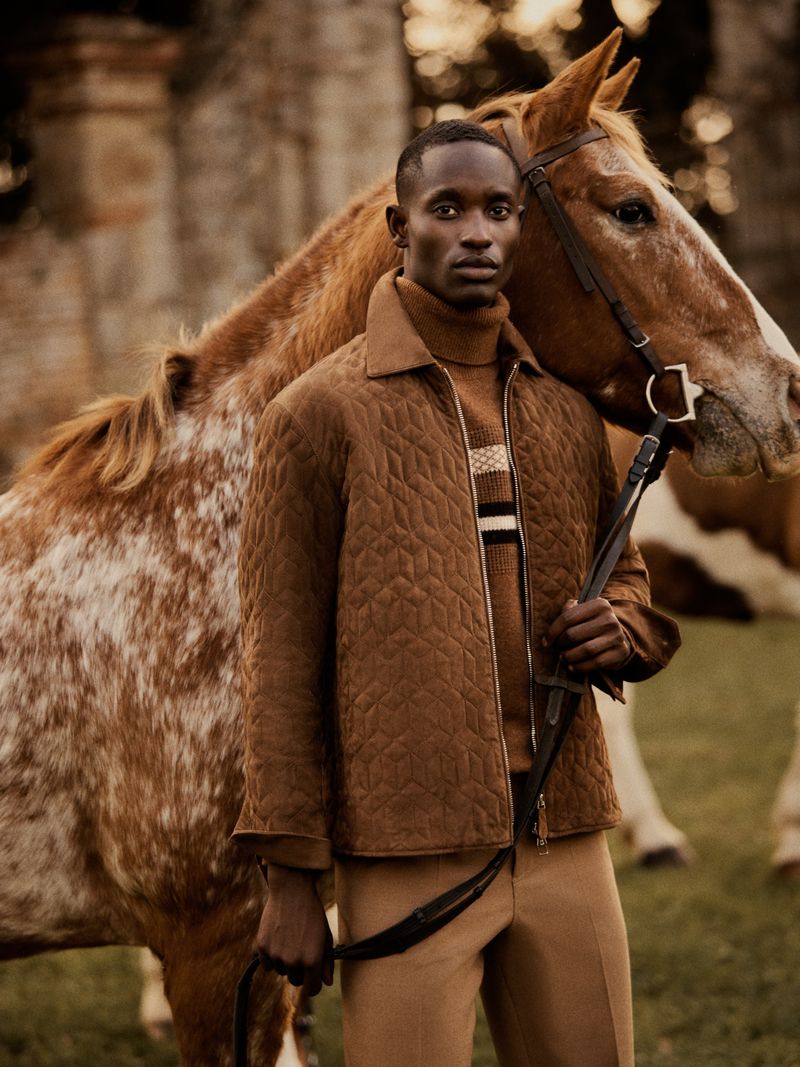 Fashion shot of a Black male model in a brown quilted jacket and tan pants, standing next to a light brown and white spotted horse.