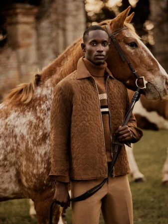 Fashion shot of a Black male model in a brown quilted jacket and tan pants, standing next to a light brown and white spotted horse.