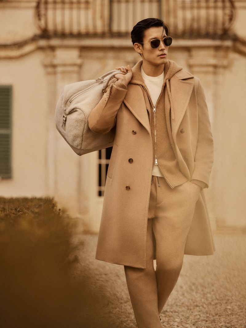 Adult male fashion model in coat walks outdoors with duffel bag, wearing sunglasses and casual separates in matching neutral tones.