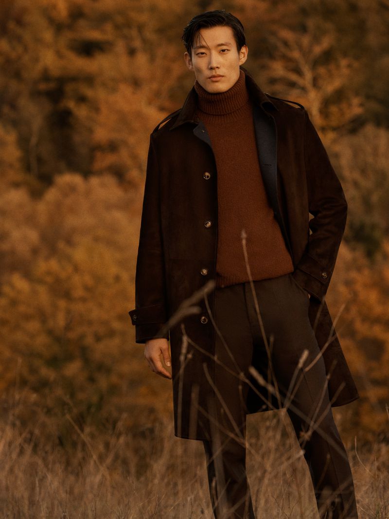 Man in brown coat and turtleneck sweater stands in a field with autumn foliage, looking directly at the camera with a serious expression.