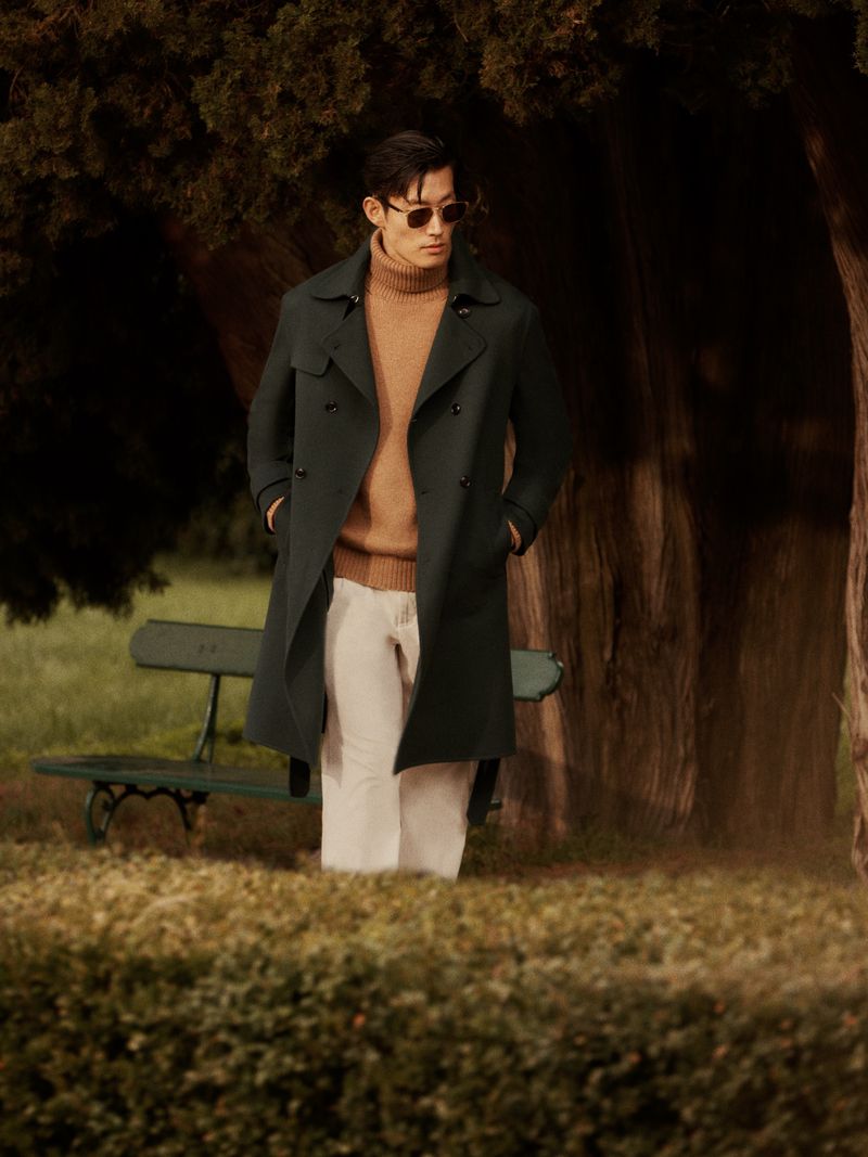 Man in sunglasses poses in trench coat, sweater, and slacks in a park-like setting with trees and bench for a fashion editorial.