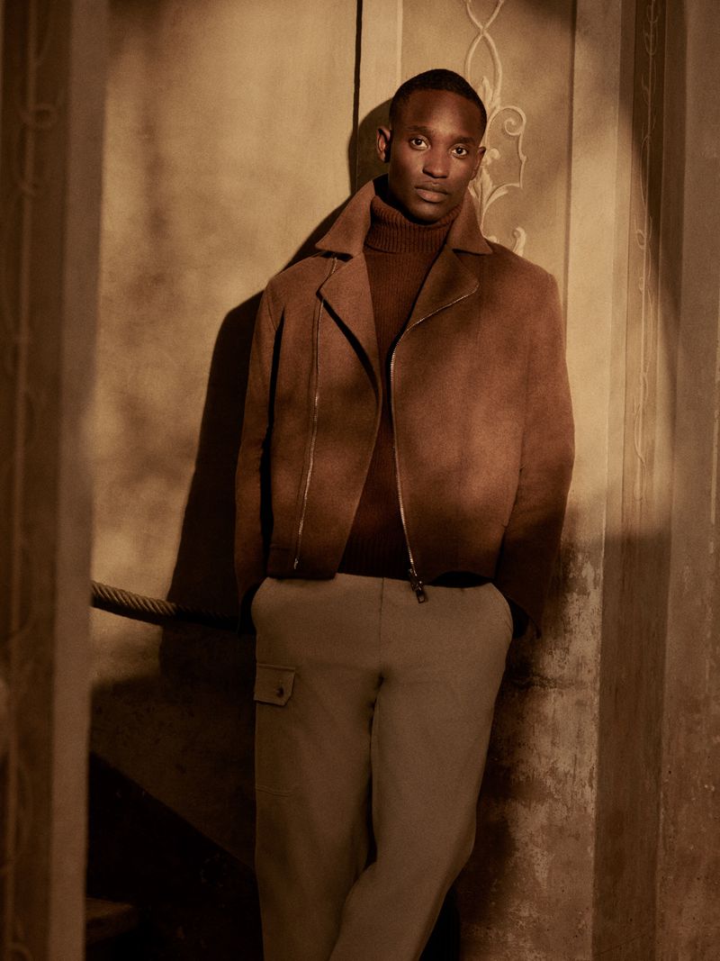 Black model standing in a brown jacket and turtleneck against an architectural wall for Brett Johnson Fall Winter 2025.