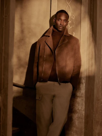 Black model standing in a brown jacket and turtleneck against an architectural wall for Brett Johnson Fall Winter 2025.