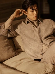 East Asian male model wearing a beige textured shirt seated on a cushioned sofa in warm lighting