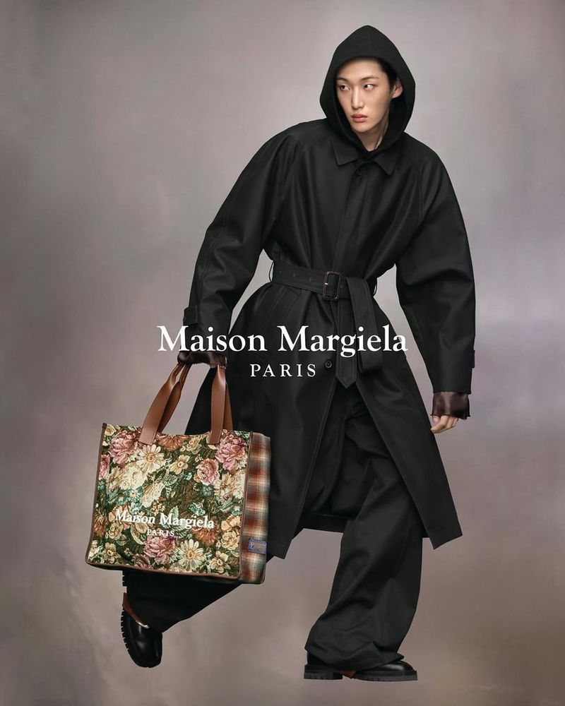 Editorial fashion shot: hooded Asian model in oversized black coat with floral tote bag, stands against soft gradient background.