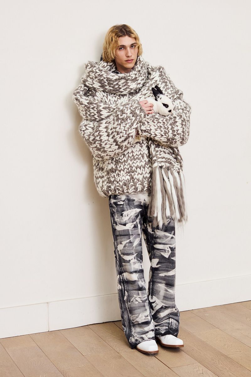 An adult male poses in an oversized knit sweater, printed pants, and white shoes while holding a toy rabbit, shot in a studio.