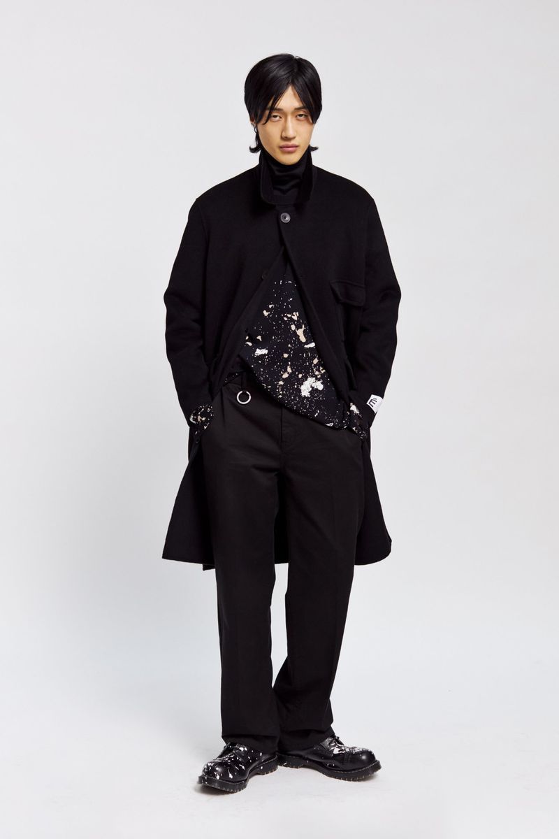 Full-length portrait of an Asian adult male in a black coat, patterned shirt, and trousers, posed against a neutral background in studio lighting.