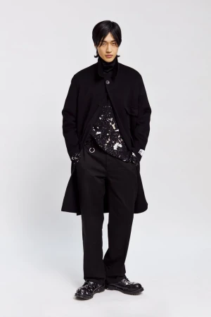 Full-length portrait of an Asian adult male in a black coat, patterned shirt, and trousers, posed against a neutral background in studio lighting.