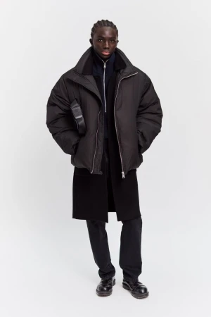 Male fashion model in dark puffer jacket, zip-up shirt, trousers, and leather shoes in a full-length studio product shot on white.