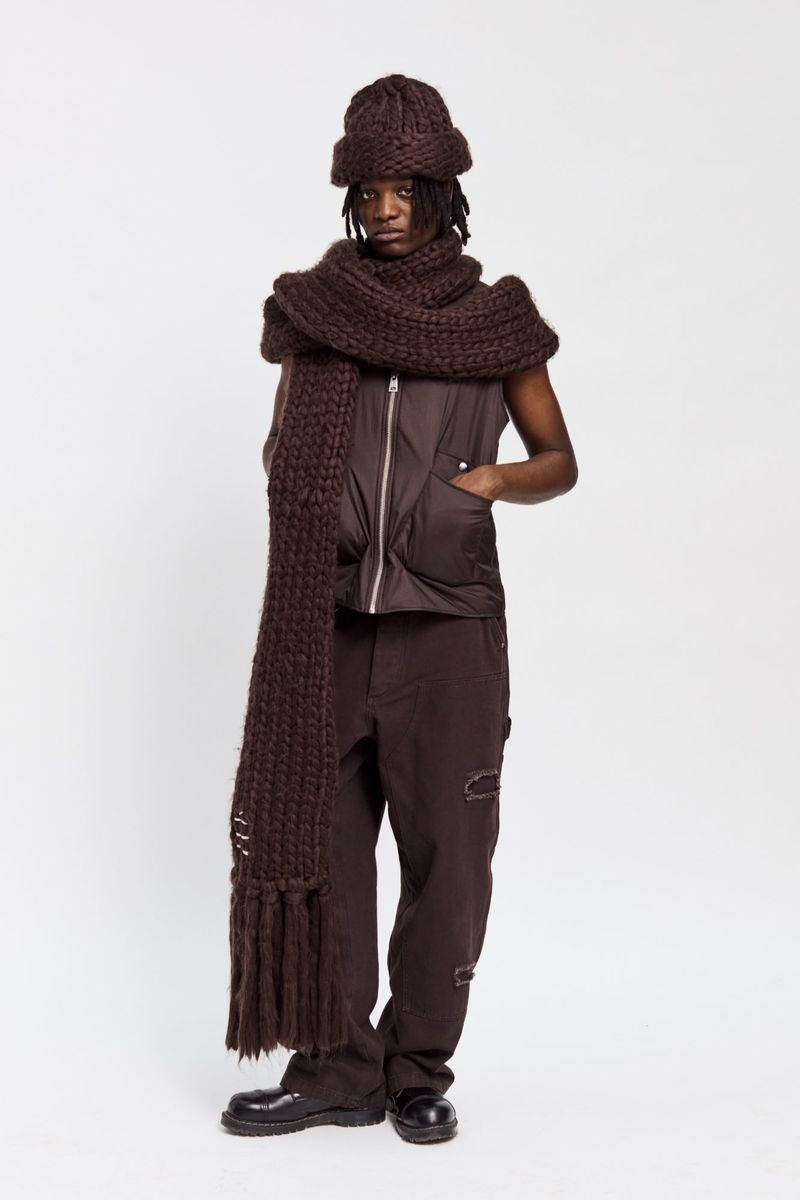 Full shot of adult male in brown knit hat, scarf, sleeveless puffer vest, pants, and black shoes in a studio with a white backdrop.