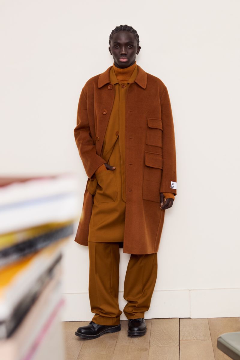 A male model wearing a brown overcoat and matching trousers standing against a plain white wall for Études Studio F/W 25.