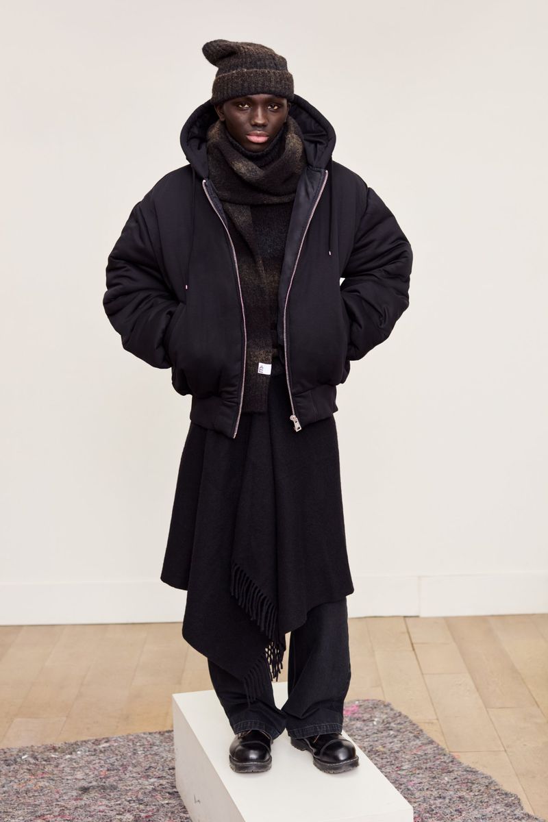 Studio shot of an adult male wearing a black hooded jacket, dark scarf, knit hat, and skirt-like garment over trousers, standing on a white platform.