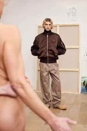 Fashion shot in studio with a blonde-haired man wearing a brown bomber jacket, tan pants, and tan shoes standing near an abstract art canvas.