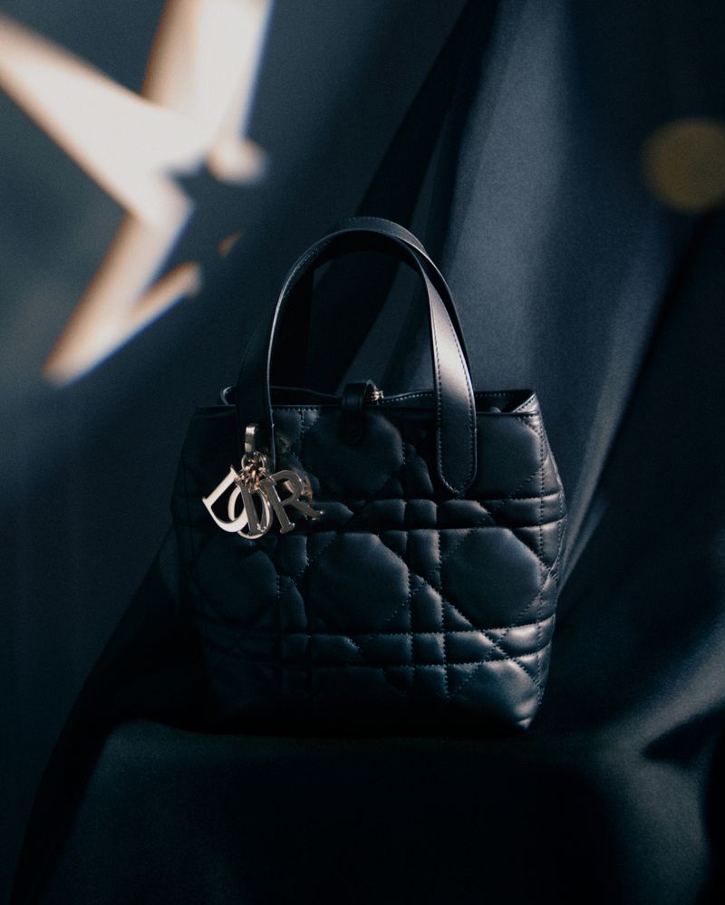 Close-up shot of a black, quilted leather Dior Lady D-Joy handbag with silver logo charms against a blurred blue background.
