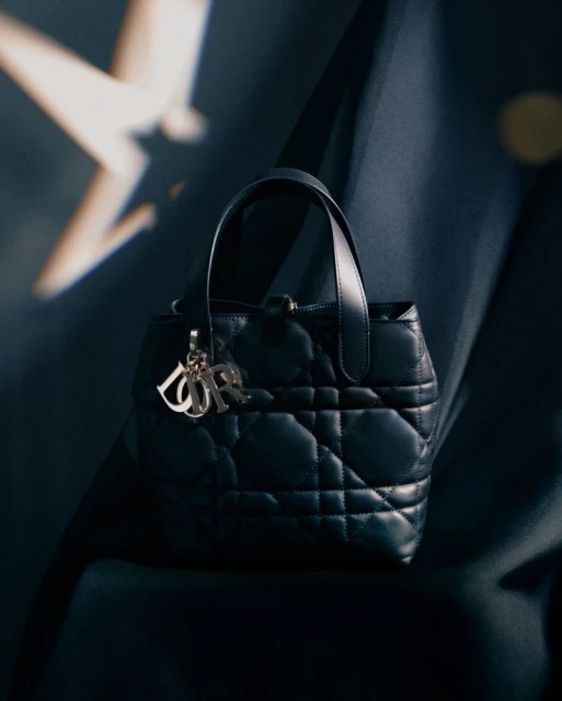 Close-up shot of a black, quilted leather Dior Lady D-Joy handbag with silver logo charms against a blurred blue background.