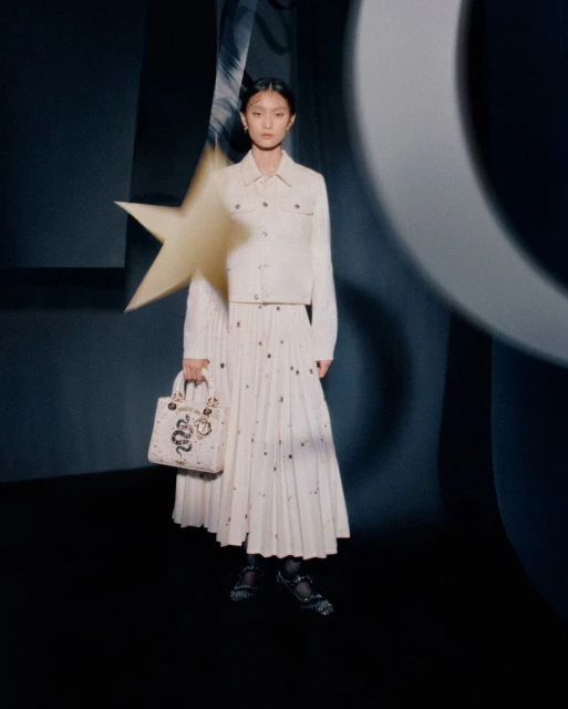 Fashion portrait of a young woman in an off-white jacket, pleated skirt, and black shoes, holding a Lady Dior handbag in a studio setting.